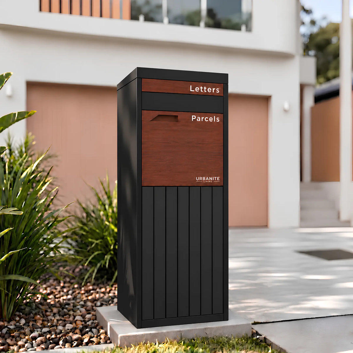 Urbanite Living Corella Extra Tall freestanding parcel letterbox in black with dark wood look metal front panels beside a driveway at a home. 