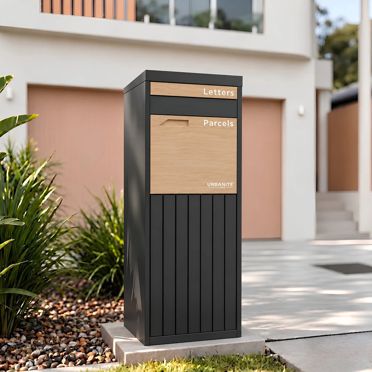 Urbanite Living Corella Extra Tall freestanding parcel letterbox in charcoal with light wood look metal front panels beside a driveway at a home.