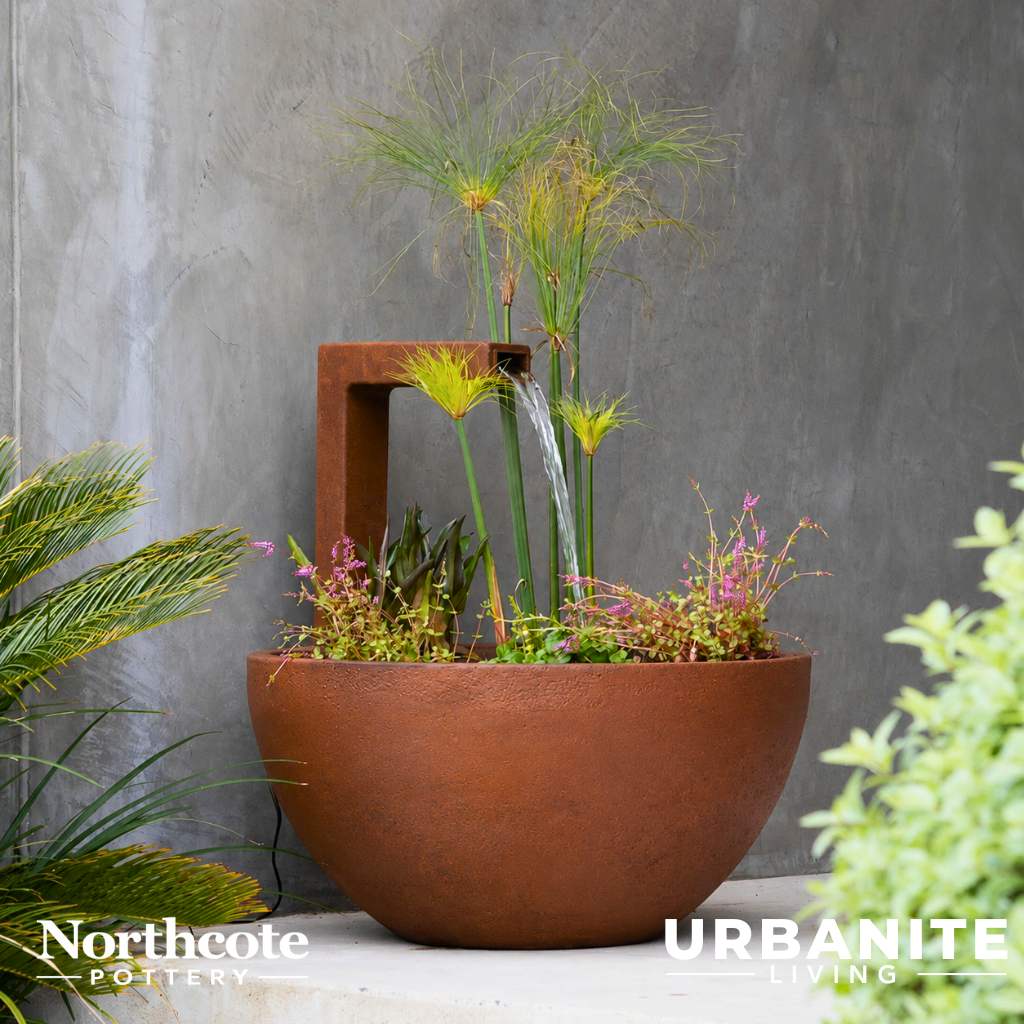 Iron Rust coloured patio pond water feature with aquatic plants and flowing water sitting on a raw concrete floor with a plastered grey wall behind.