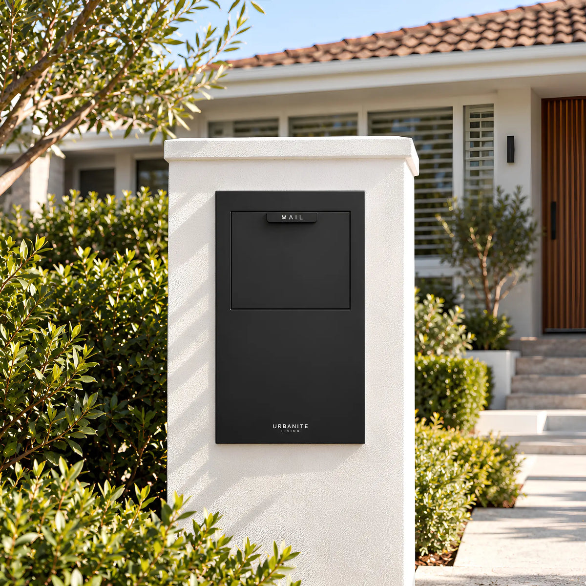 Urbanite Living Wren parcel letterbox in black, set into a white rendered pillar in a front garden.  